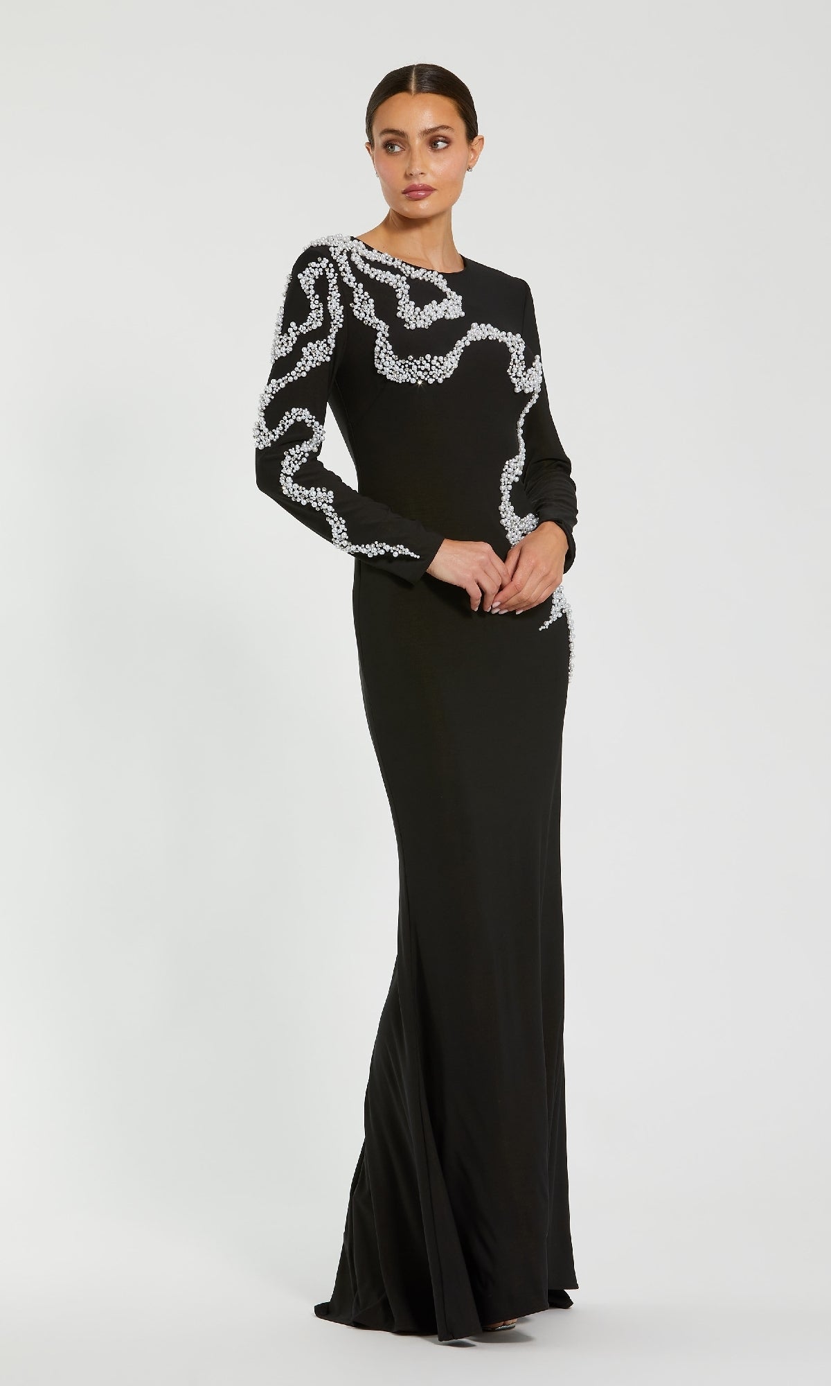 A woman stands against a plain light background, wearing the Mac Duggal 44086 long formal dress—a striking black jersey evening gown with white embellished patterns on one sleeve and across the bodice.